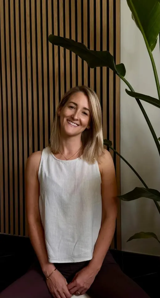 Annalena lächelt freundlich in die Kamera, stehendes Porträt vor einer Holzlamellenwand mit Grünpflanze