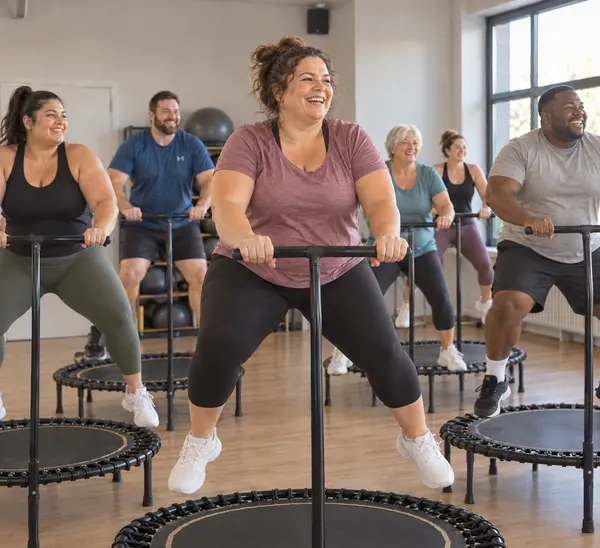 Lächelnde Frau springt entspannt auf einem Mini-Trampolin mit Haltestange, umgeben von einer diversen Gruppe – Stimmungsbild für den Kurs Curvy Jumping